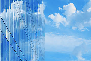 A modern glass office builidng reflects blue sky and clouds indicating environmental architecture or a business intent to reduce greenhouse gas emissions.